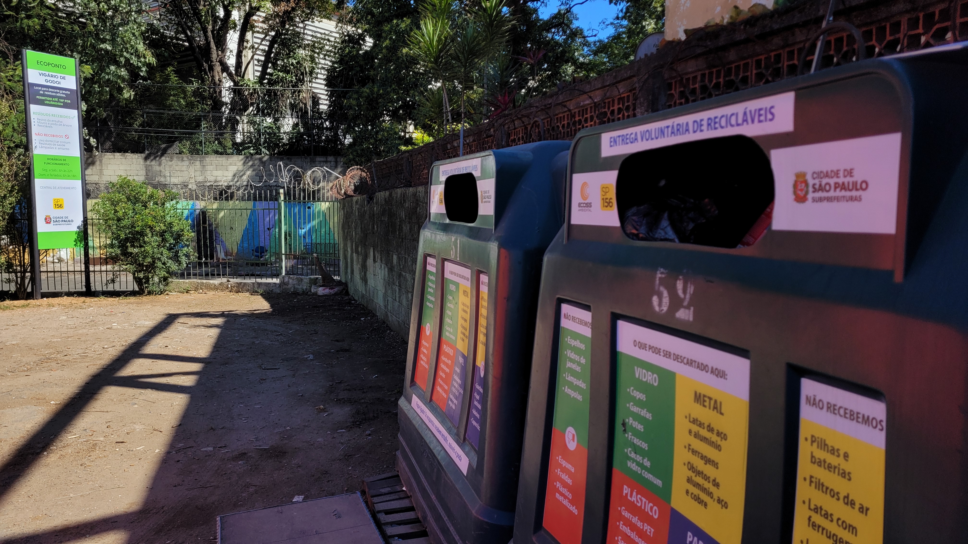 A imagem mostra um ecoponto de reciclagem identificado como "Ecoponto Vigário Godói", localizado na cidade de São Paulo. No primeiro plano, há contentores verdes de coleta seletiva com adesivos coloridos informando quais tipos de materiais podem ser descartados, como vidro, plástico e metal, além de uma lista de itens não permitidos. O local é destinado à entrega voluntária de recicláveis. Ao fundo, há um portão metálico com um muro de concreto, parcialmente coberto por grafites, além de vegetação e uma placa informativa com regras e orientações sobre o descarte adequado de resíduos. O ambiente externo está ensolarado, projetando sombras no chão de terra.